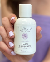 5 Circle Skin Care Naked Oil Cleanser in their hand, with a purple towel draped behind them in a softly lit indoor setting.