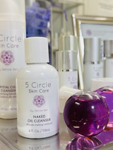 A bottle of 5 Circle Skin Care Naked Oil Cleanser in the nude color, shown alongside other skincare products in a reflective, premium setting.